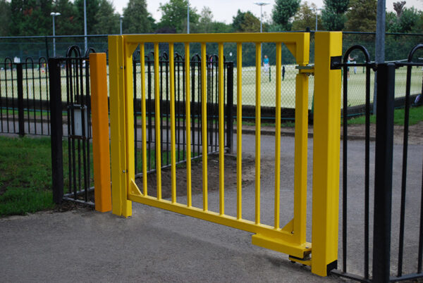 Yellow metal pedestrian gate at leisure centre with self-closing gate hinge on bottom hinge and pivoting top hinge